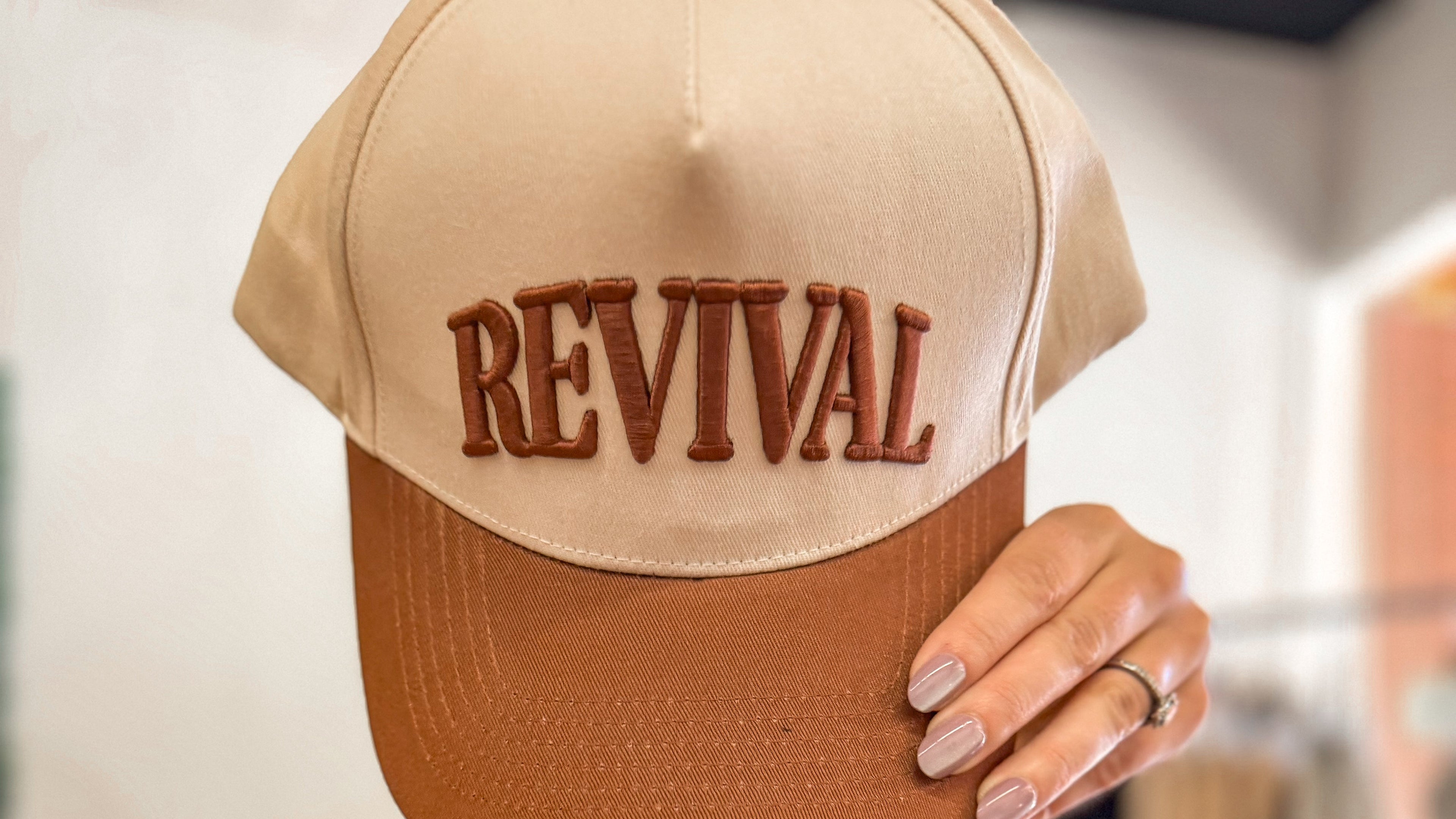 Beige and brown cap with 'REVIVAL' text held by a person wearing a green dress.